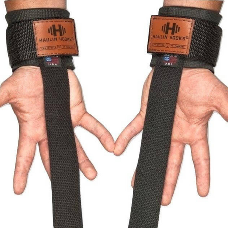 Hands wearing "HAULIN' HOOKS 'STRAP 650'" weightlifting straps with brown patches and U.S. flag tags, featuring neoprene wrist padding for comfort, proudly made in the USA.