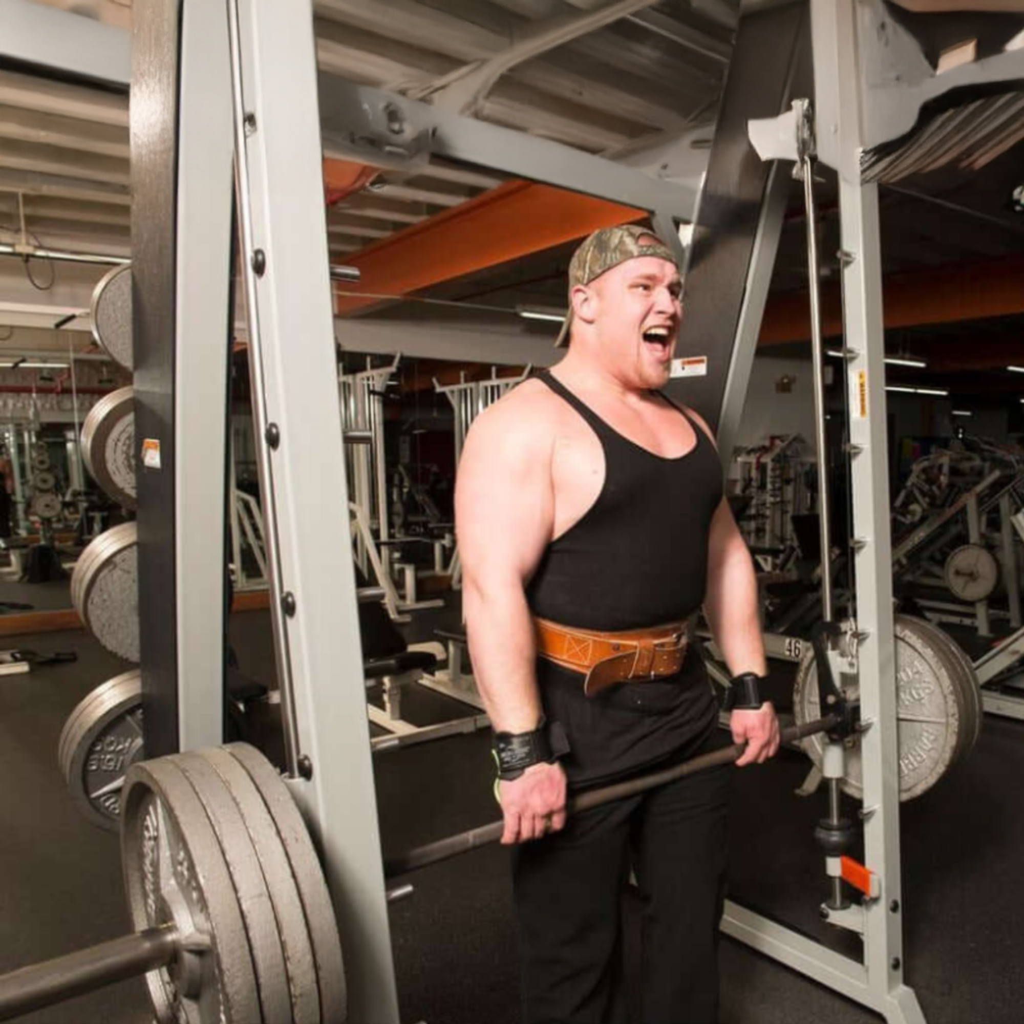 A muscular man at the gym confidently lifts heavy weights using HAULIN HOOKS ONLYS, wearing a black tank top and lifting belt, and smiles.