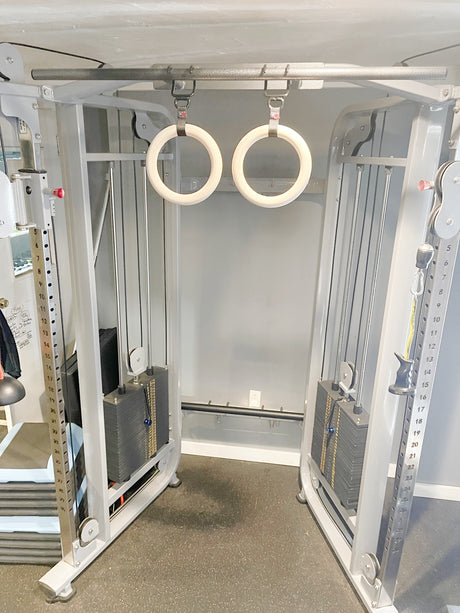 A cable machine features GYMNASTICS RINGS Pull up Handles Roman Ring Pull Up Grips with wooden handles suspended in the center, set within a modern gym environment.