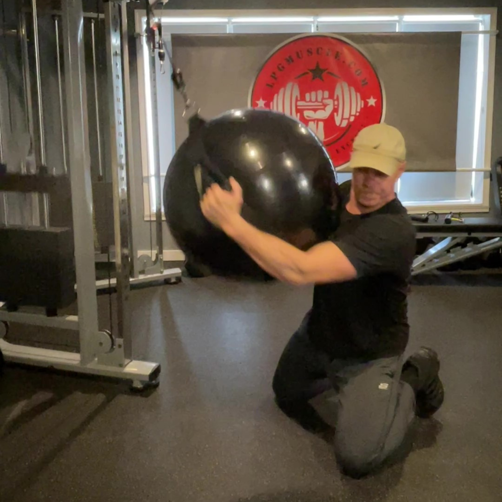 Man kneels in a gym, using PHYSIO TWIST Swiss Ball Handle Straps for Cable Machine Core to perform twists with a resistance ball, building explosive core power.