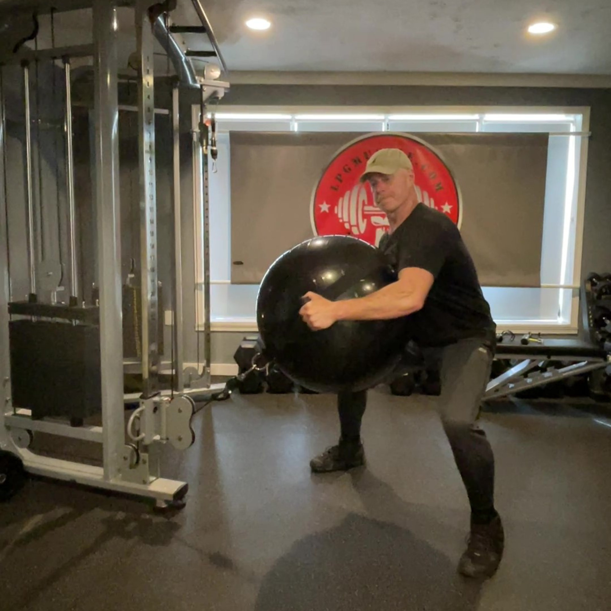 A man in the gym uses PHYSIO TWIST Swiss Ball Handle Straps for Cable Machine Core to squat with a large exercise ball, highlighting intense core engagement and explosive strength, surrounded by gym equipment.