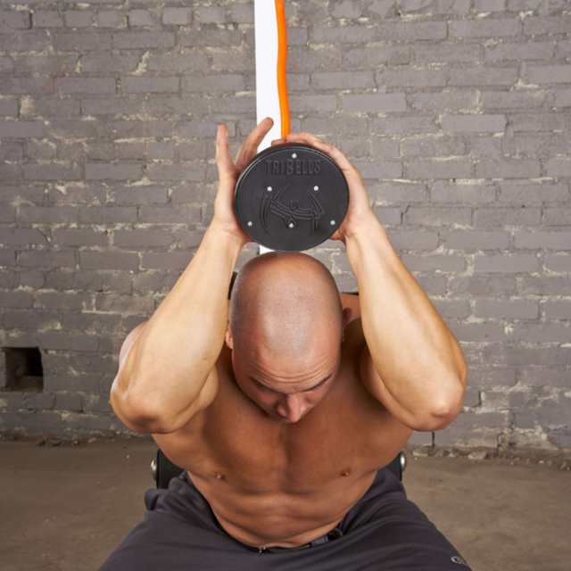 A shirtless man demonstrates a tricep extension in front of a gray brick wall using the TRIBELLS 6" Palms Grip Tricep attachment, which destroys dumbbells!.