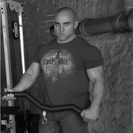 A muscular man holding a workout bar in a gym, wearing a "Grip Freak" t-shirt, facing forward.