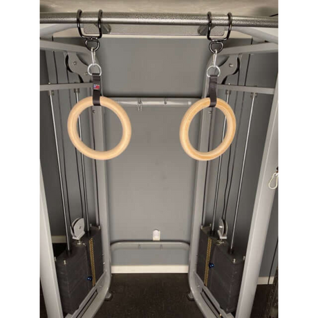 A pair of wooden gymnastic rings hangs from a cable machine in a fitness gym.
