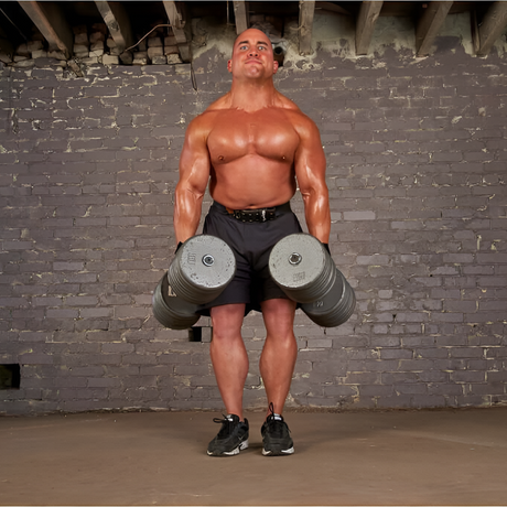 A muscular man stands holding large dumbbells at his sides in a gym with a gray brick wall.