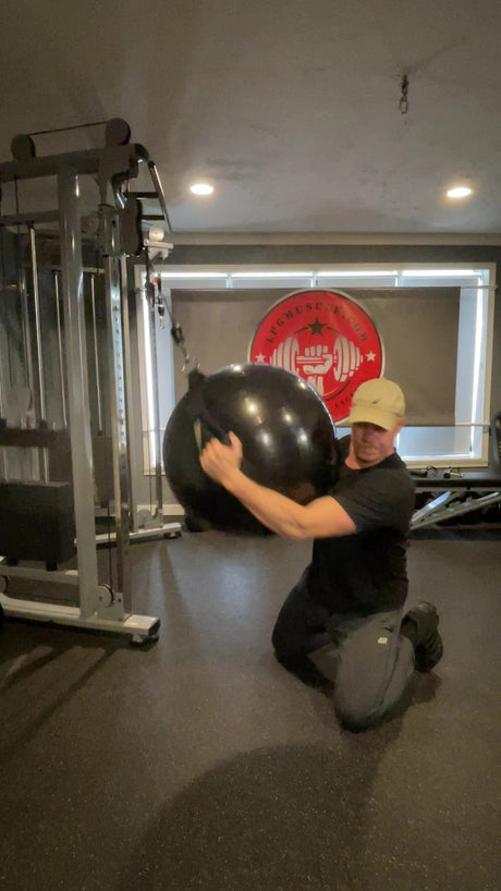 A man kneels in a gym, pulling a cable machine using a large exercise ball.