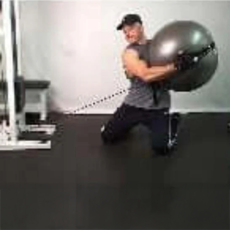Man kneeling on gym floor, twisting while holding an exercise ball connected to a cable machine.