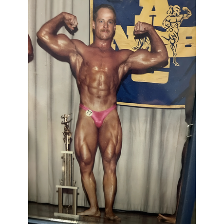 A male bodybuilder in pink trunks flexes on stage with a trophy and a bodybuilding banner behind him.
