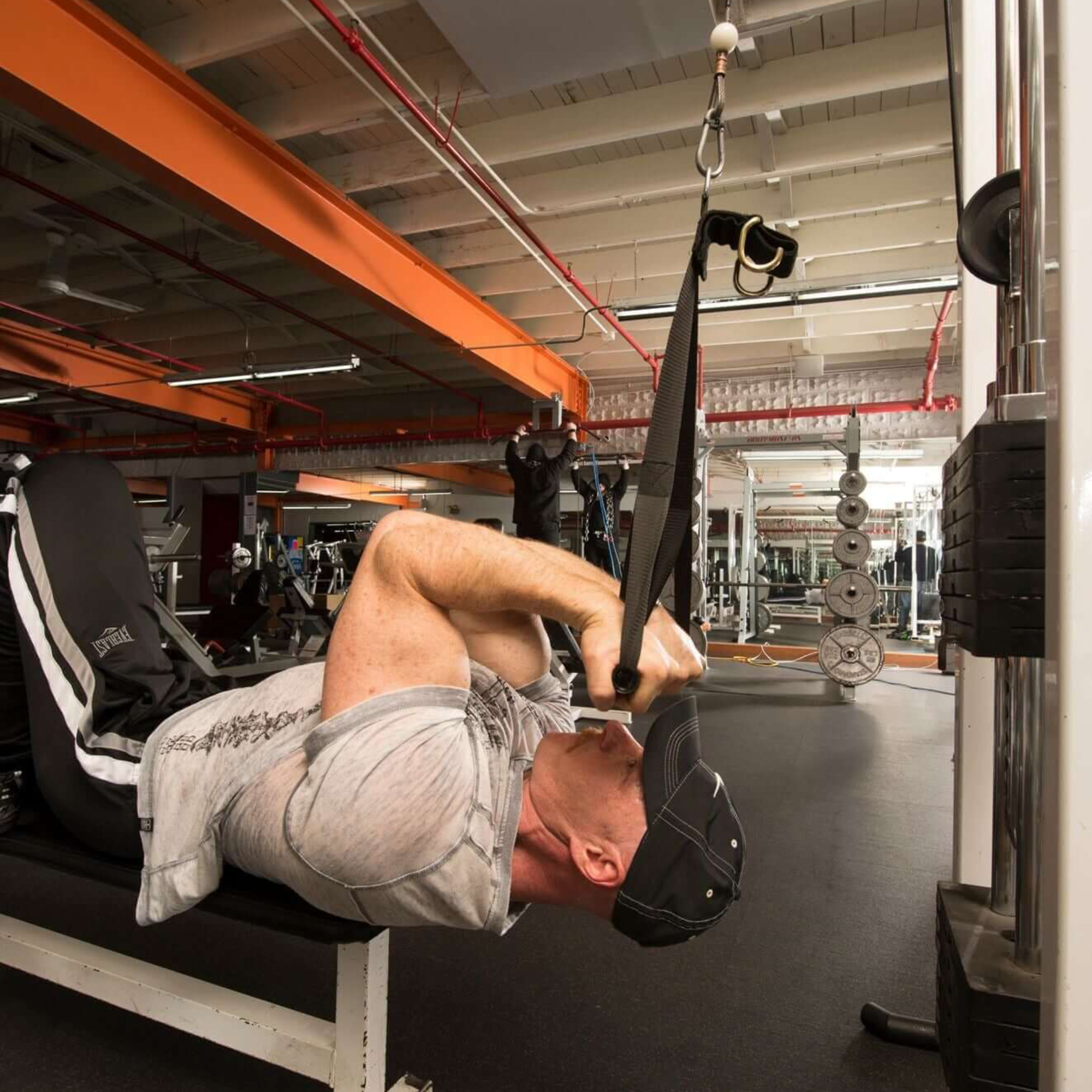 A man lies on a gym bench doing triceps extensions with the DELT-BELT Handle Straps for Cable Machine Upright Rows, while a barbell and other multi-exercise handles nearby offer added workout variety.