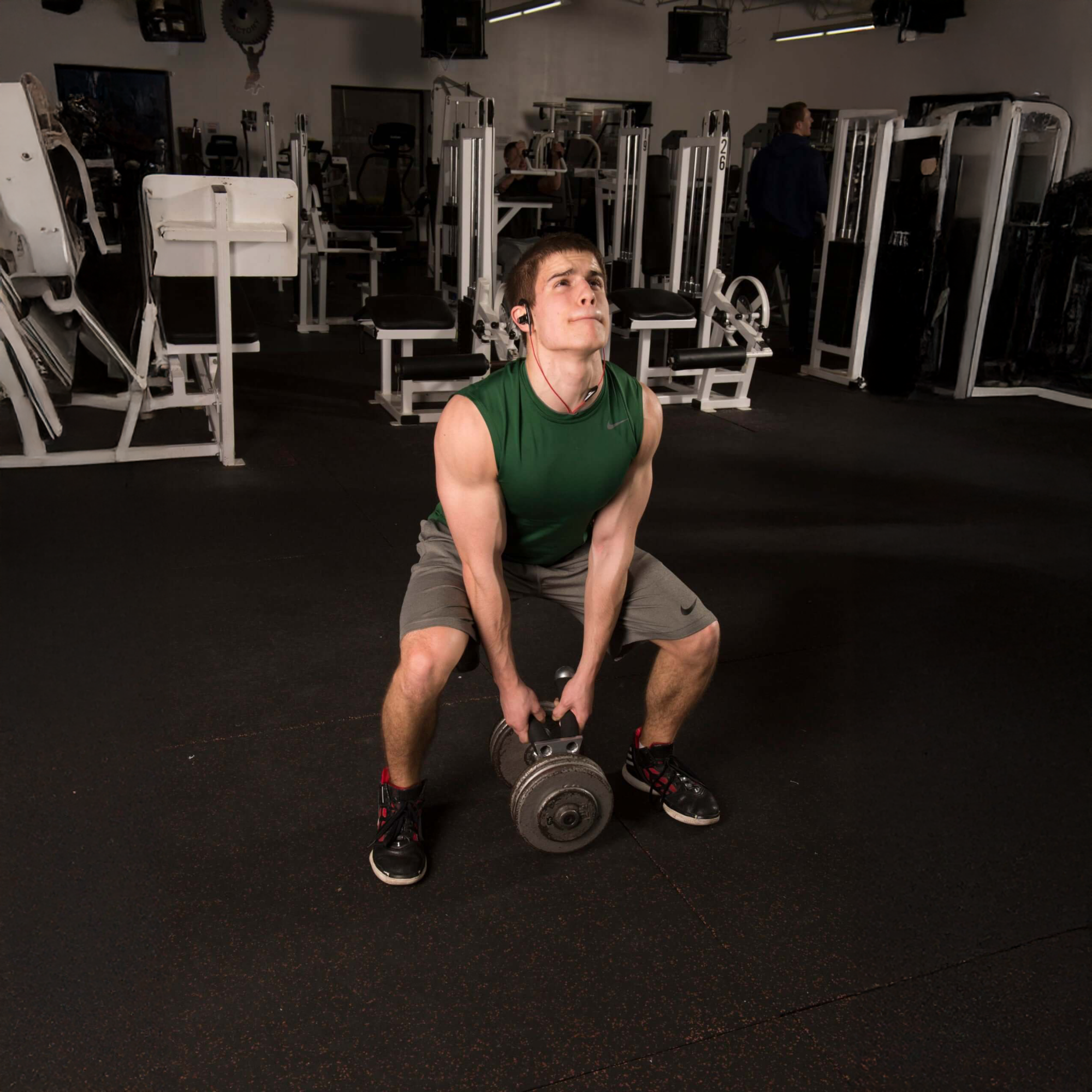 Wearing a green sleeveless shirt and shorts, a man at the gym uses the EZ SQUAT Dumbbell Gym Equipment for Legs Squat Handles to lift a heavy weight from the floor.