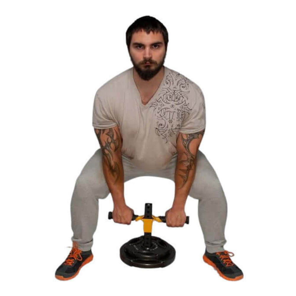 A tattooed, bearded man squats in his home gym, using the EZ SQUAT Handles System Squat Machine Squat Attachment with an Olympic weight plate. Dressed in a beige shirt and gray pants, he exudes strength and focus in his workout regimen.