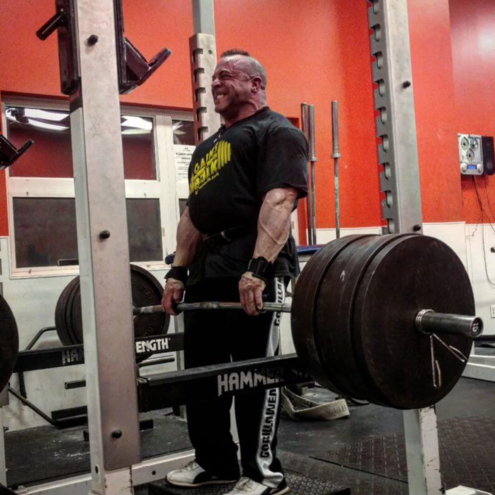 In a gym, a man uses HAULIN HOOKS 'HARDCORE' 2 in 1 Weightlifting Hook Straps Combination to deadlift heavy weights on a barbell, displaying muscular effort amidst surrounding gym equipment.
