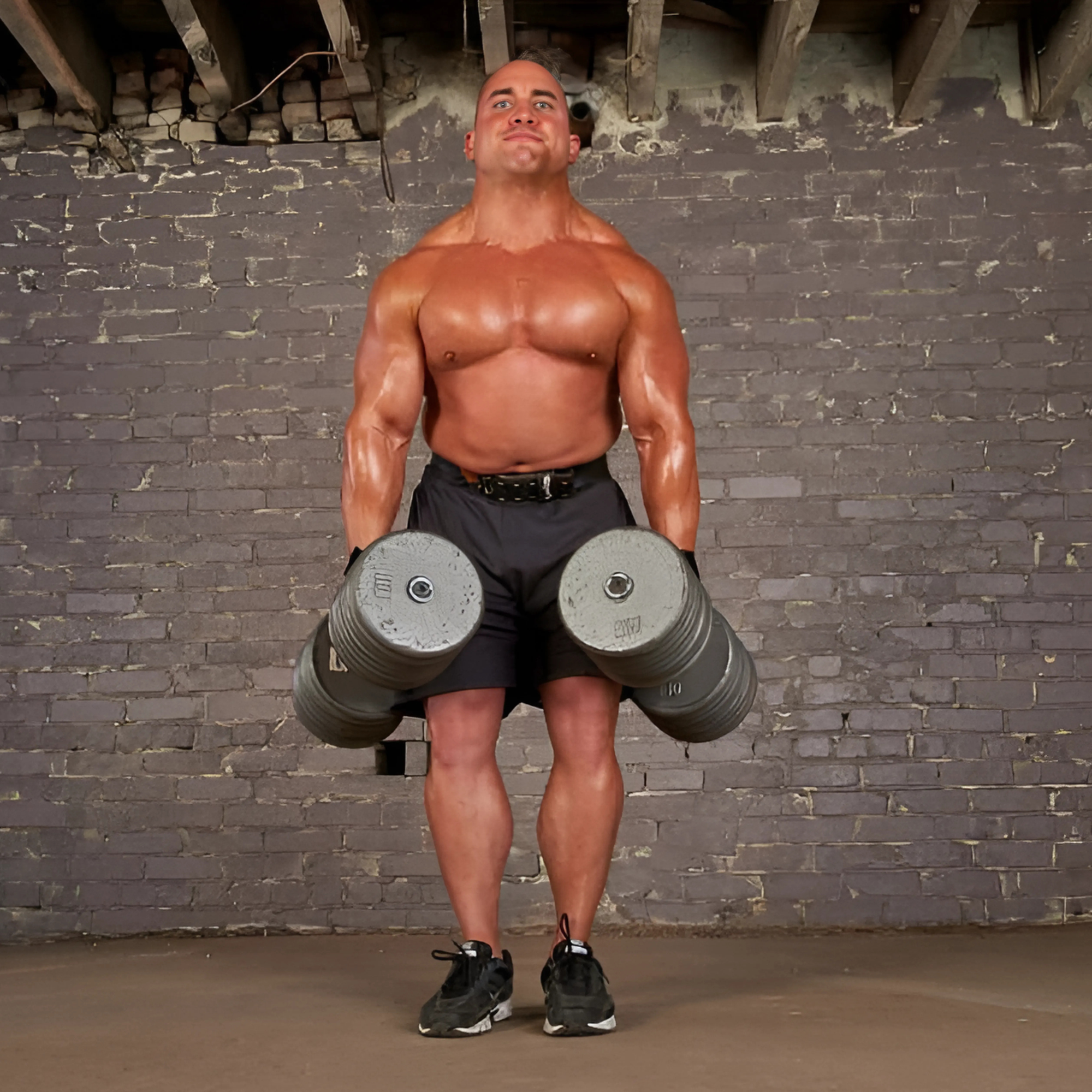 Muscular man in shorts lifts heavy dumbbells with HAULIN HOOKS 'ORIGINALS' Hooks for Weightlifting Extreme, gym brick wall in background.