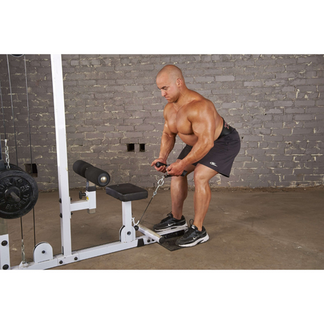 A man performs a cable squat in the gym, using the USA-made HOG LEGS 'Back Pac' LEGIT Cable Machine Attachments Set against a gray brick wall.