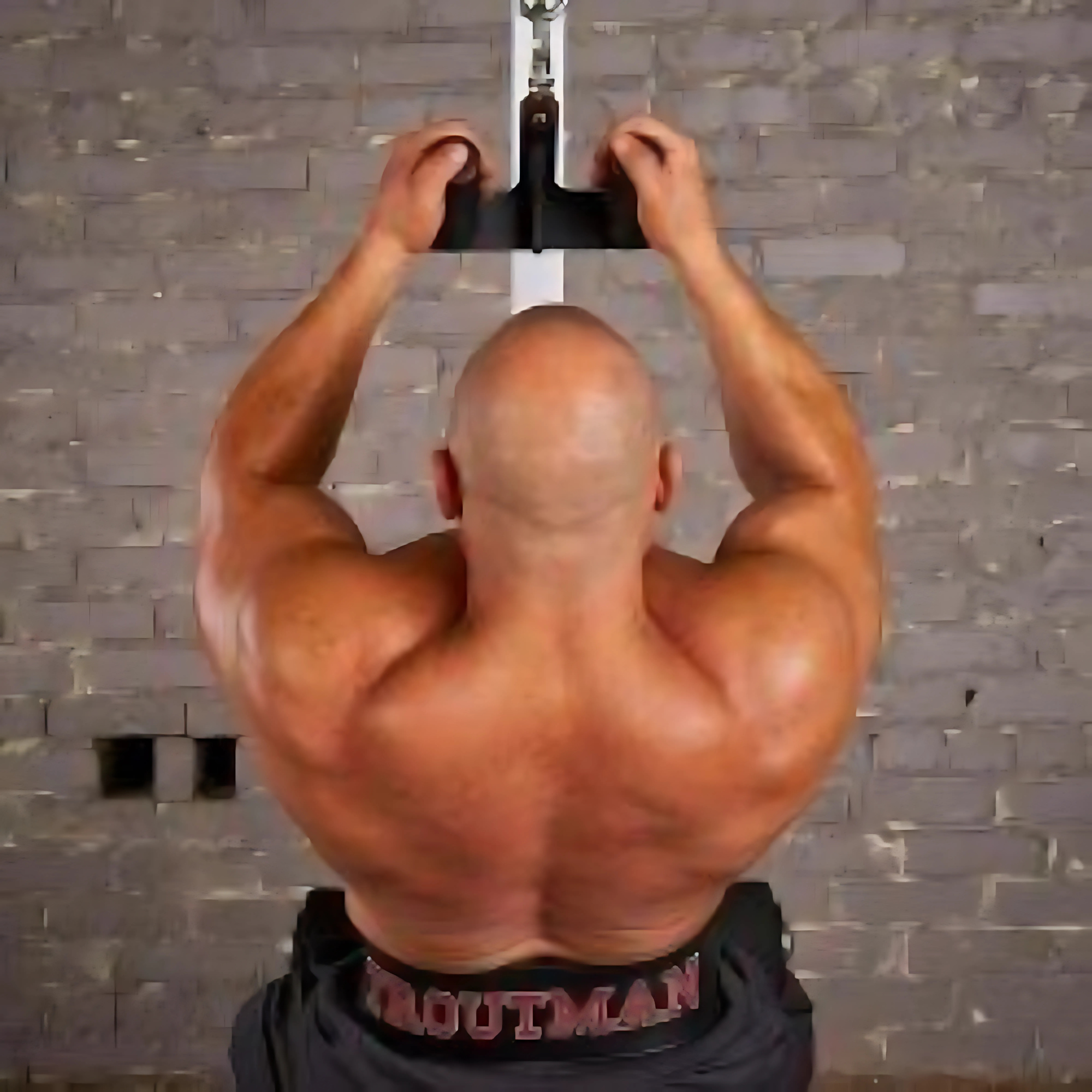 A muscular man seen from behind grips a HOG LEGS 'Back Pac' LEGIT Cable Machine Attachments Set against a brick wall background.