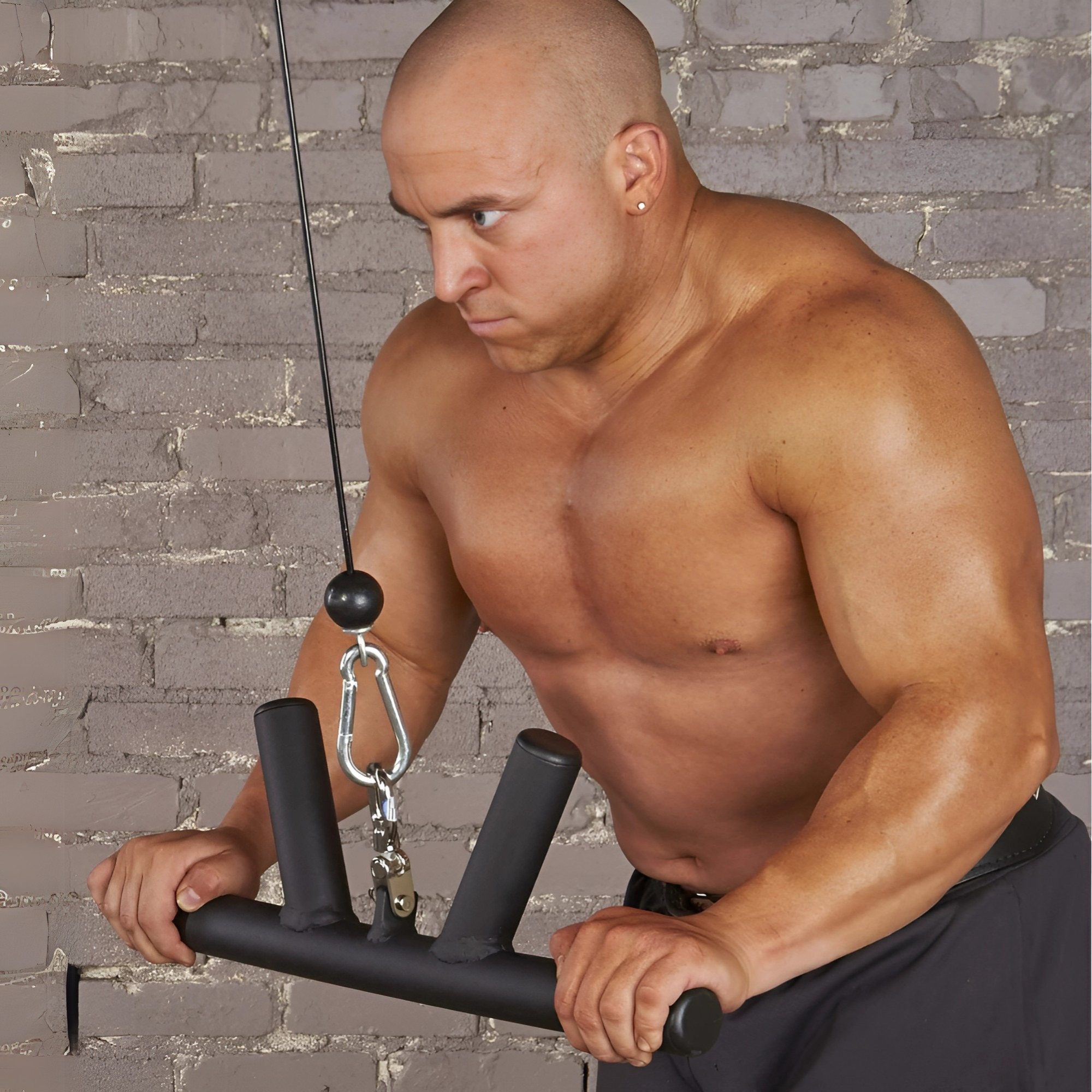 A muscular man intensively works his triceps and forearms in the gym using the HOG LEGS 'Boar Tusk' Fat Grip Curling Bar for cable exercises, with a gray brick wall as the backdrop.