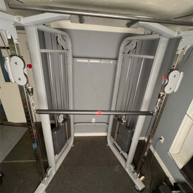 A home gym with HOG LEGS Dual Cable Bar Attachments sits against a gray wall, offering versatile workout options with adjustable weight stacks and dual pulleys.