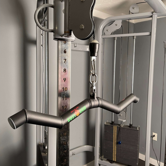 A close-up of the HOG LEGS 'Feral Hog' Arm Workouts with Cables Bar set, featuring a V-bar attachment for forearms, biceps, and triceps. The fat cable bars and weight stack are prominently visible in the gray room.