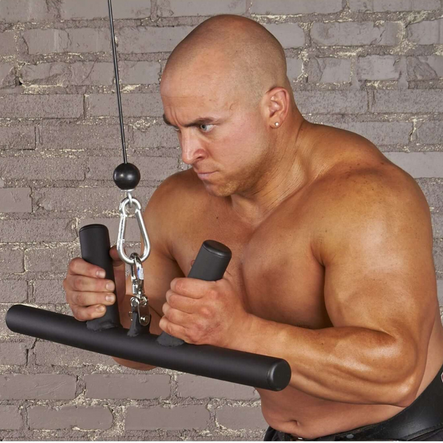 A muscular man uses the HOG LEGS 'Feral Hog' cable bar set for forearm, bicep, and tricep exercises against a brick wall. The gym's commercial-grade equipment ensures each workout meets his highest standards.