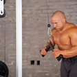 A muscular man uses the HOG LEGS 'Feral Hog' Arm Workouts with Cables Bar set on a cable machine for triceps exercises in a gym with a gray brick wall.