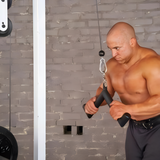 A muscular man uses the HOG LEGS 'Feral Hog' Arm Workouts with Cables Bar set on a cable machine for triceps exercises in a gym with a gray brick wall.