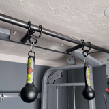 Two black "HOG LEGS 'Fist Full' Pullup Handles & Tricep Attachment" dangle from a metal pull-up bar in a home gym, enhancing grip strength for challenging workouts.