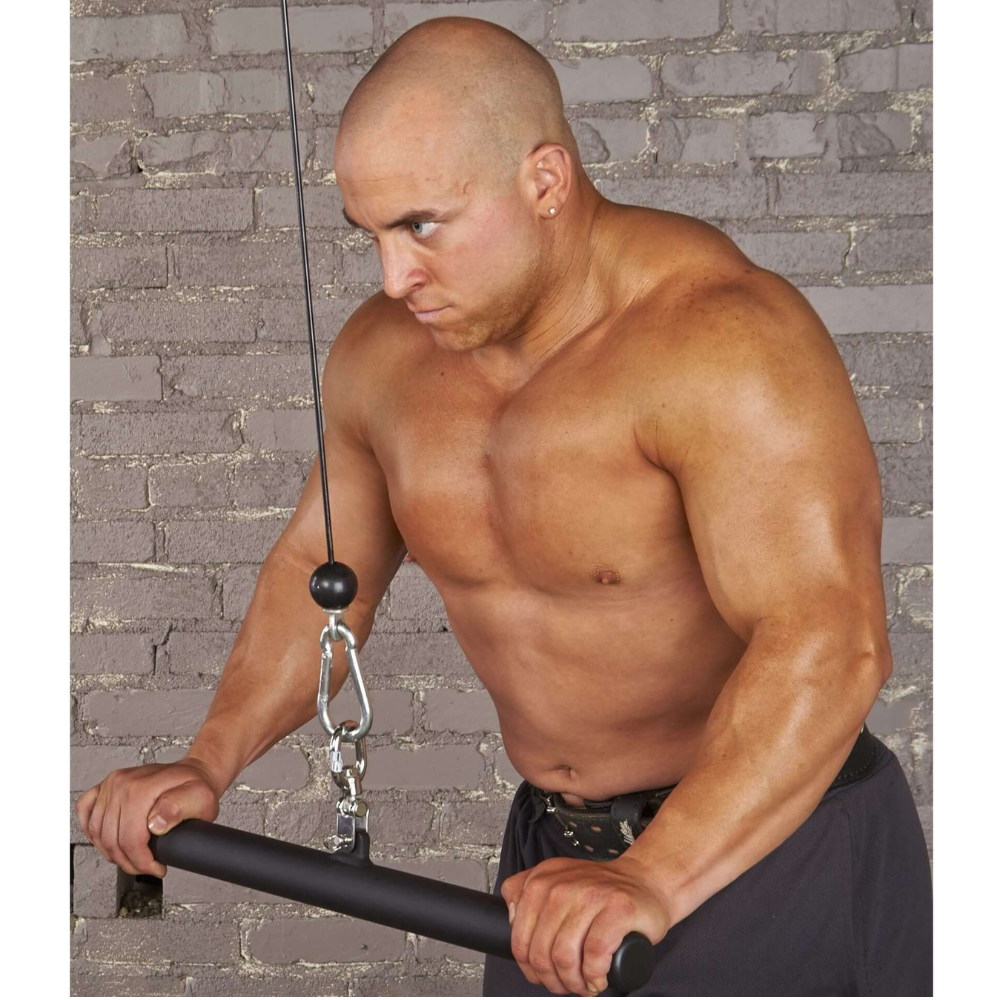 A man with a shaved head, gripping the HOG LEGS 'Ham Hock' FAT Cable Curl Bar Extreme, works out on a cable machine. He leans forward with a serious expression against a brick wall backdrop.
