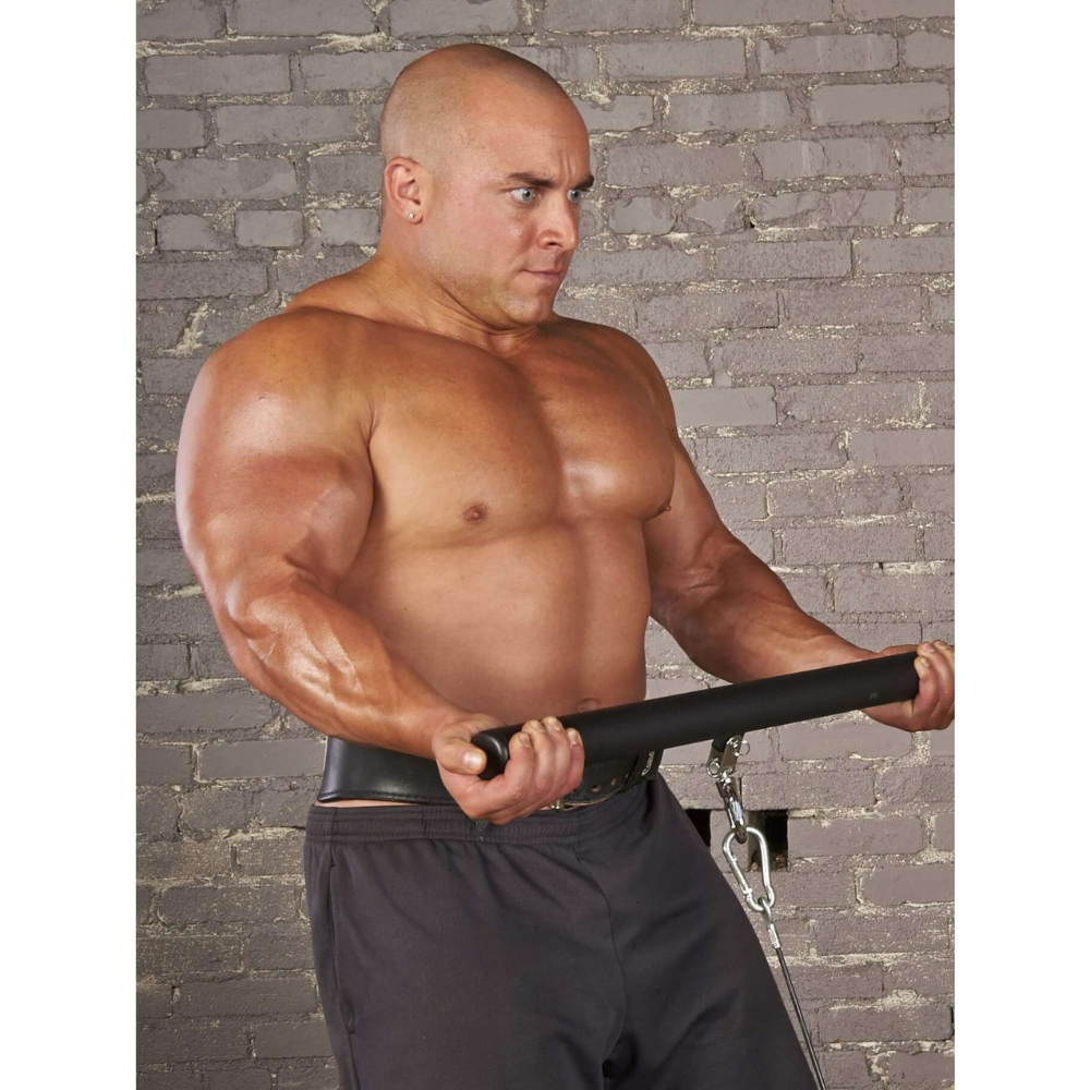 A bald, muscular man lifts a HOG LEGS 'Ham Hock' FAT Cable Curl Bar Extreme indoors, wearing black shorts against a gray brick wall.