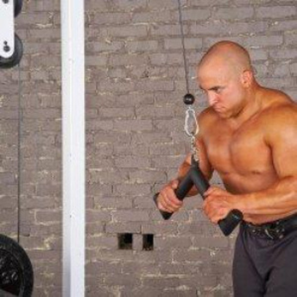 A man works out on a cable machine at the gym, focusing intently on triceps extensions using the HOG LEGS 'Pork Chop' Killer Triceps Workout Cable Bar, in front of a gray brick wall.
