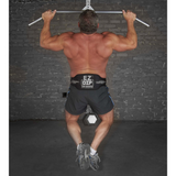 A man, clad in shorts and sneakers, performs a pull-up at the gym. He showcases strength and determination with the LOADING PIN for Weights plus Dip Belt with chain HOOK strapped around his waist, facing a brick wall.