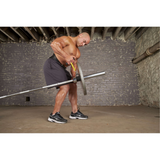 In a dimly lit basement, a man in black shorts and sneakers focuses on his workout, executing bent-over rows using the PULL FORCE 'Single' Landmine Attachment for Barbell Handle.