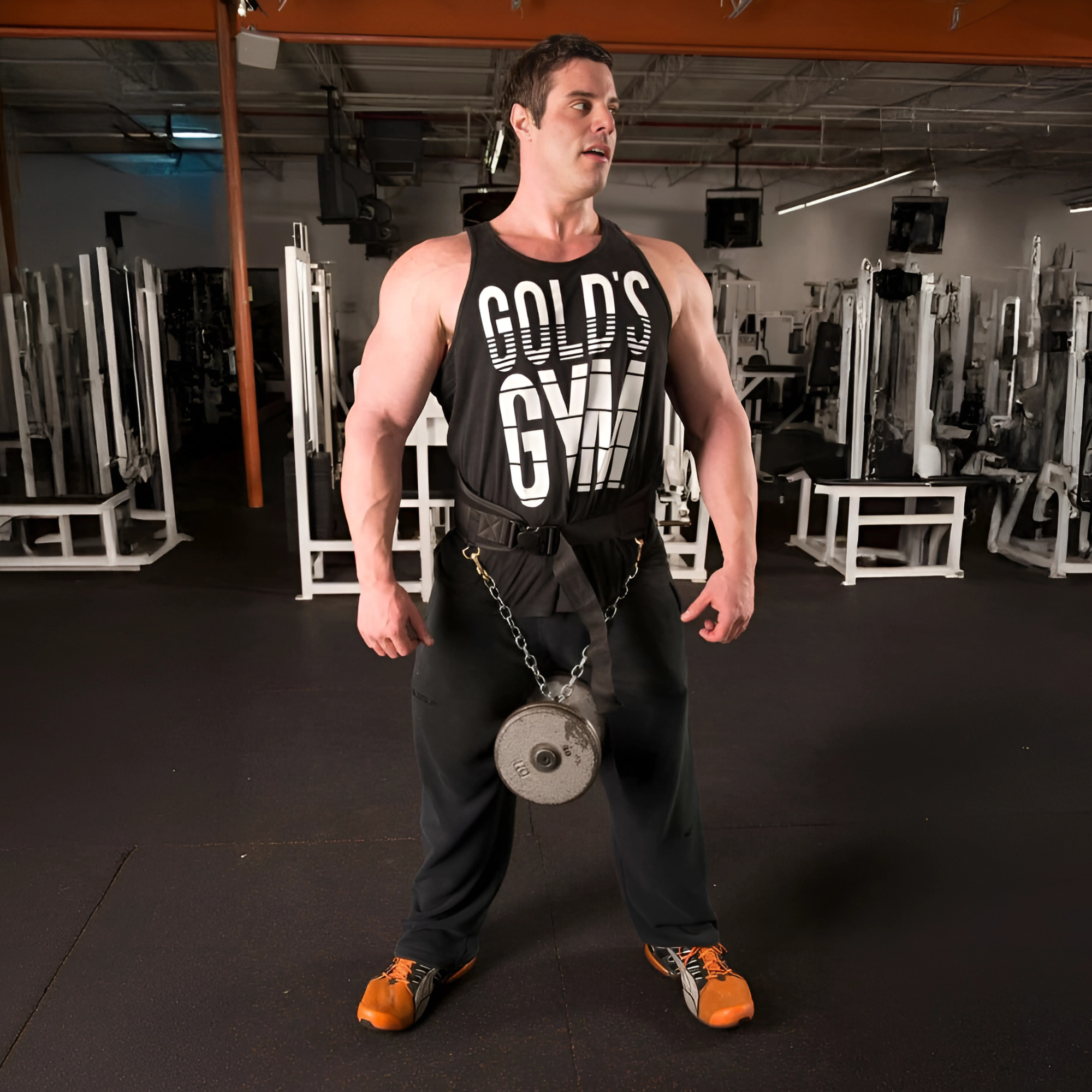 A man in a Gold’s Gym tank top stands in a gym, using a Patented DIPPING BELT HOOK for Dip Belt with Chain, with a weight plate attached around his waist.