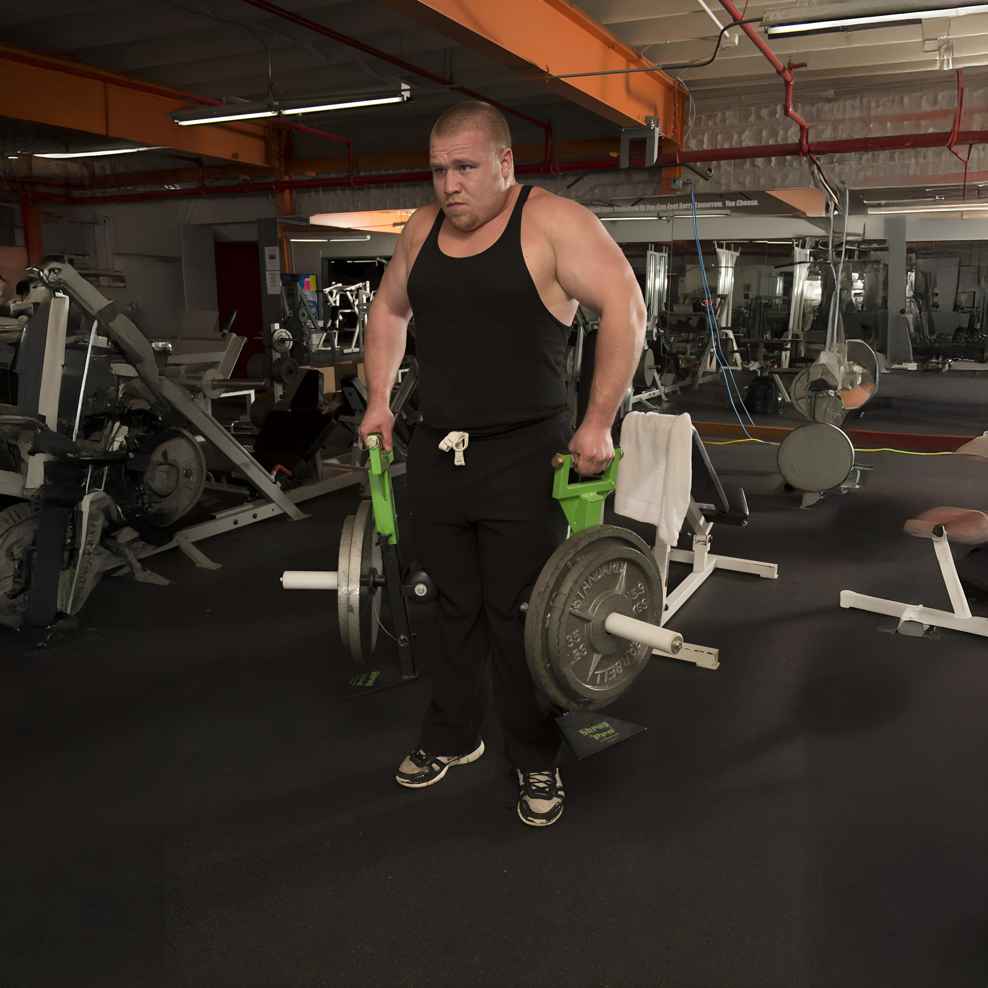 A man at the gym wearing a black tank top holds heavy weights using the SHRUG PRO Freestanding Hex Bars Trap Bar Barbell Shrug Alternative (Without Rollers), featuring a patented freestanding design and adjustable handles for maximum performance.