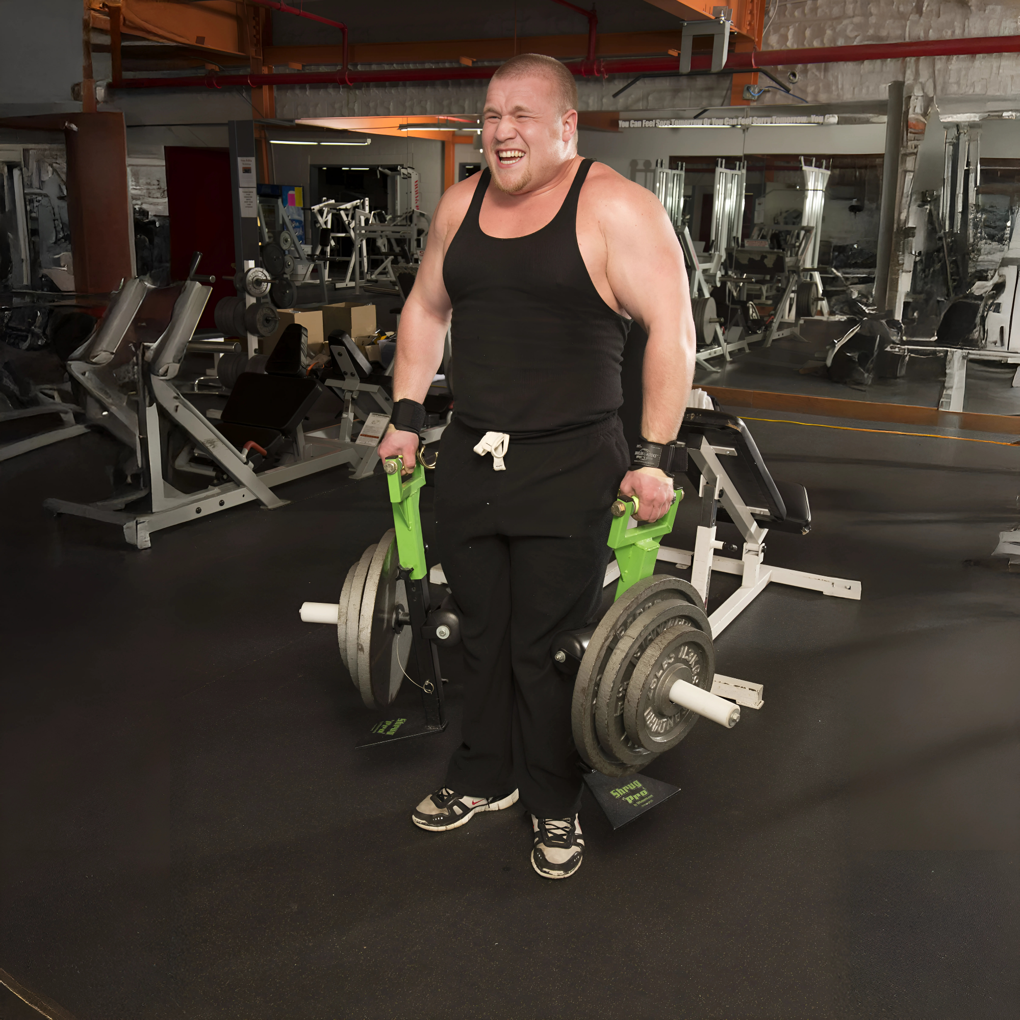 A man in a black tank top lifts heavy weights using the SHRUG PRO Freestanding Hex Bars Trap Bar Barbell Shrug Alternative—Without Rollers, straining with effort in a gym.