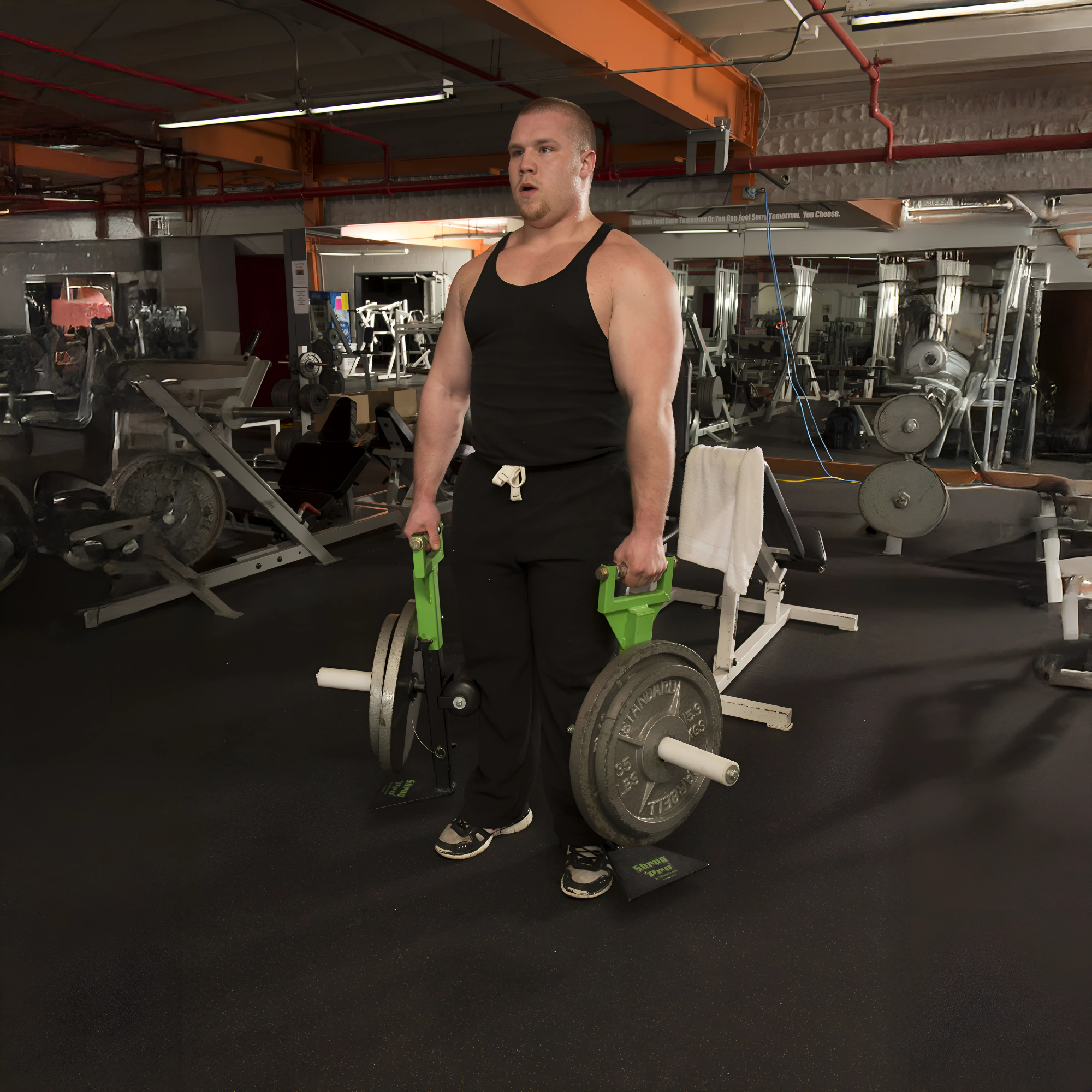 A man in black gym gear lifts weights with green handles on a SHRUG PRO Freestanding Hex Bars Trap Bar Barbell Shrug Alternative—Without Rollers, surrounded by Olympic Weight Plates in a gym.