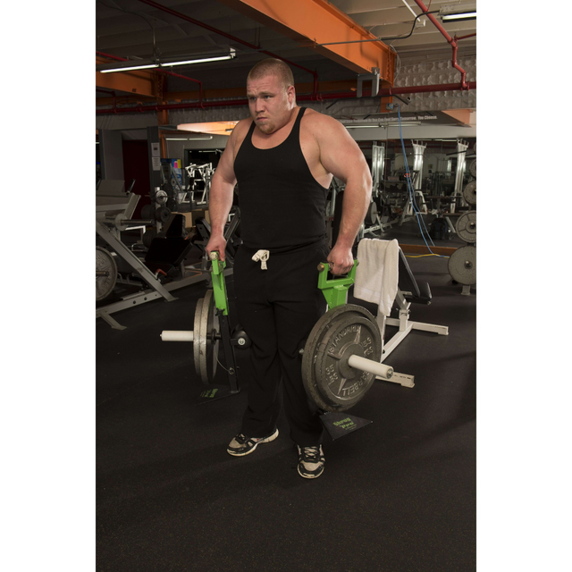 A man in a gym is using the 'SHRUG PRO' Freestanding Hex Bars Trap Bar Barbell, lifting weights with green handles, and wearing a black tank top and pants.