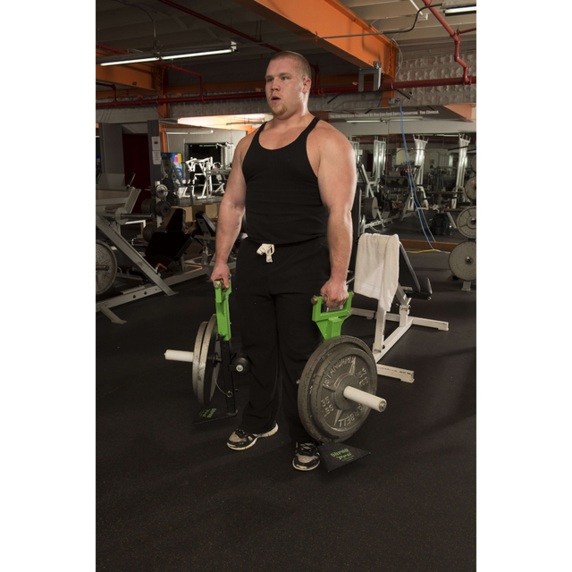 A person in a gym is lifting heavy weights with farmer's walk handles, skillfully maneuvering past gym equipment, including a 'SHRUG PRO' Freestanding Hex Bar resting nearby.
