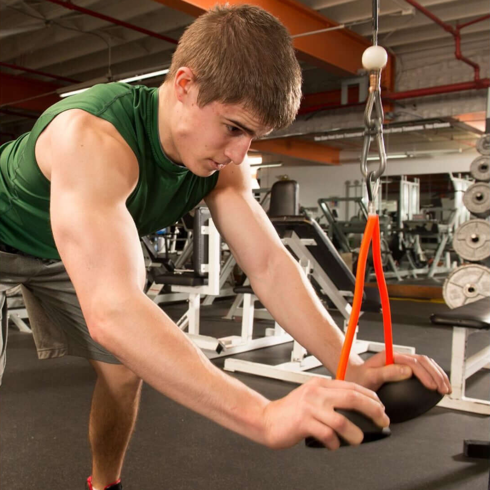 Wearing a green sleeveless shirt and gray shorts, a person confidently uses resistance bands at the gym, expertly incorporating TRIBELLS 4" 'Double' Tricep Workout with Cables Gym Handles to intensify their exercise, showcasing both strength and style.