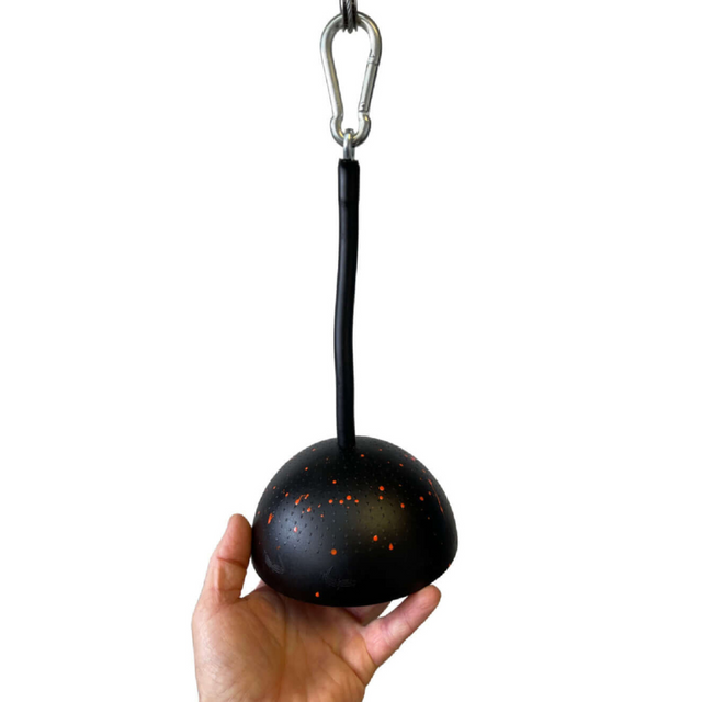 A hand holds the TRIBELLS 6" Palms Grip, a black hanging grip ball with orange speckles, attached to a hook by a short strap, perfect for enhancing tricep exercises on the cable machine.