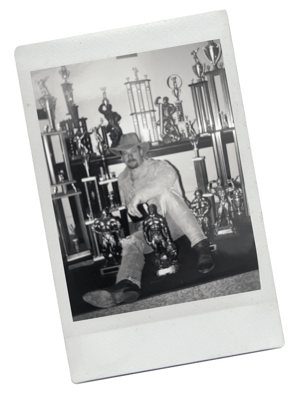 A man sits on the floor surrounded by many trophies and awards in a black and white photograph.