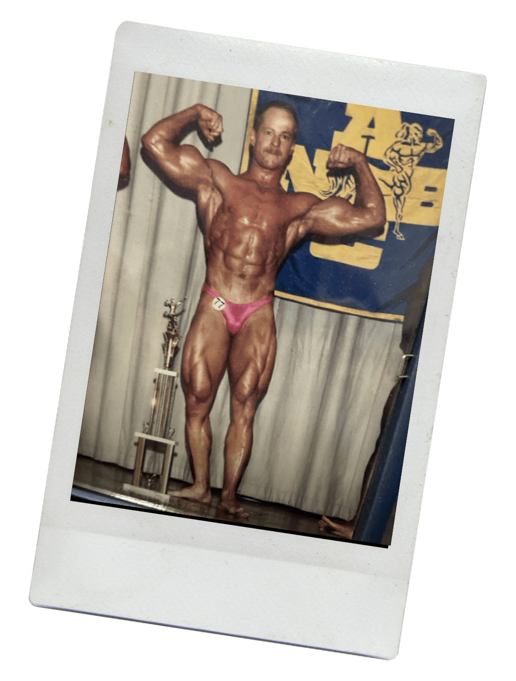 A male bodybuilder in pink briefs poses on stage with a trophy nearby and a competition banner behind him.